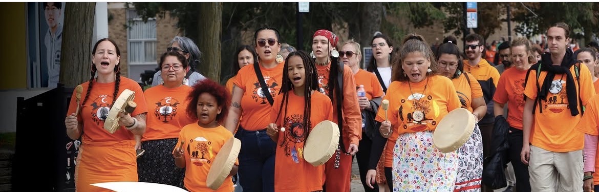 Community participants in orange shirts walking and playing hand drums